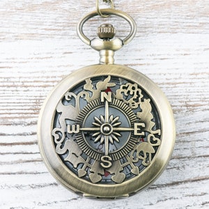 Op de afbeelding: Een messing zakhorloge met een kompasgezicht en ingewikkelde decoratieve details rond de rand. Het kompasgezicht heeft een zwarte achtergrond met een zilveren kompasnaald, een zilveren ster en de letters N, S, E en W.