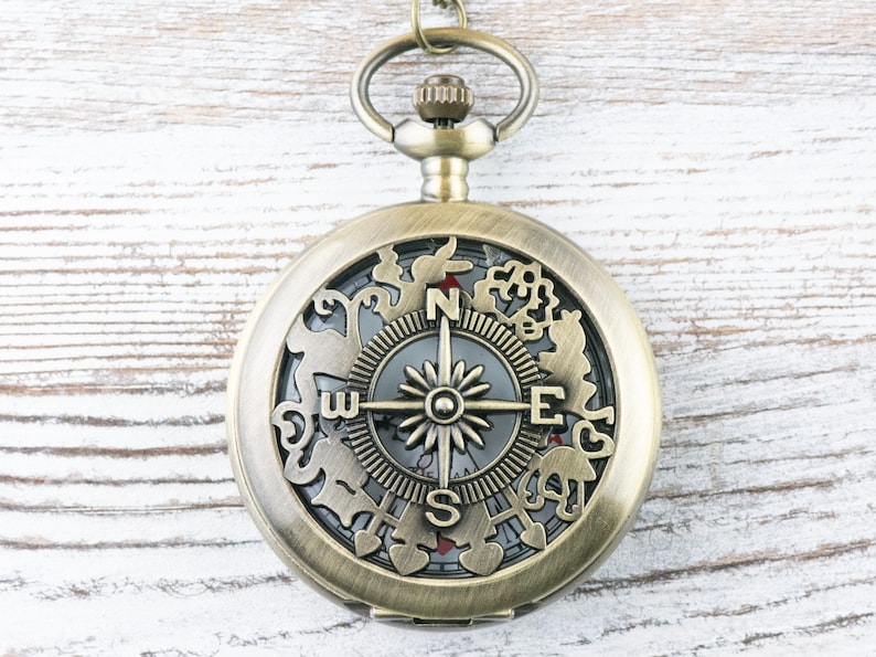 Op de afbeelding: Een messing zakhorloge met een kompasgezicht en ingewikkelde decoratieve details rond de rand. Het kompasgezicht heeft een zwarte achtergrond met een zilveren kompasnaald, een zilveren ster en de letters N, S, E en W.