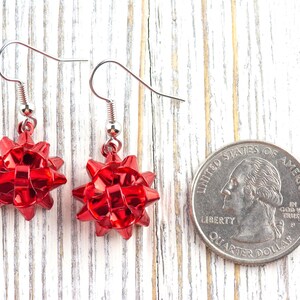 May include: A pair of red bow-shaped earrings with silver-colored hooks. The earrings are made of a shiny, reflective material. A U.S. quarter coin is next to the earrings for size comparison. The earrings are on a white wooden surface.