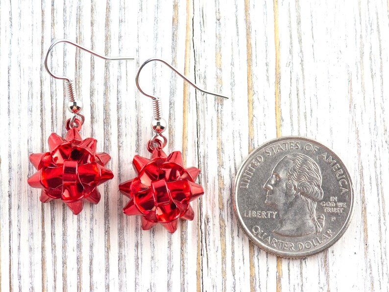 May include: A pair of red bow-shaped earrings with silver-colored hooks. The earrings are made of a shiny, reflective material. A U.S. quarter coin is next to the earrings for size comparison. The earrings are on a white wooden surface.
