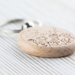 May include: A wooden keyring with the word "Faith" engraved on it, surrounded by a floral design. The keyring is light brown and has a silver metal ring.
