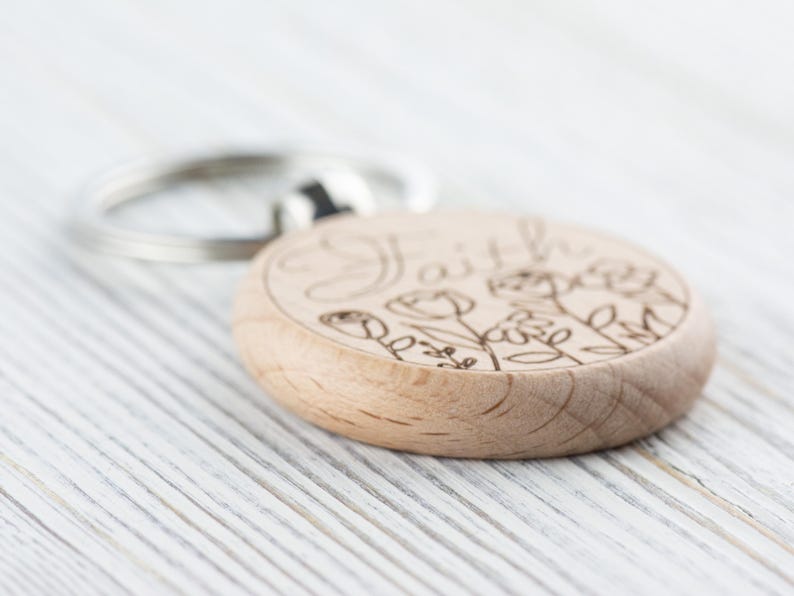 May include: A wooden keyring with the word "Faith" engraved on it, surrounded by a floral design. The keyring is light brown and has a silver metal ring.