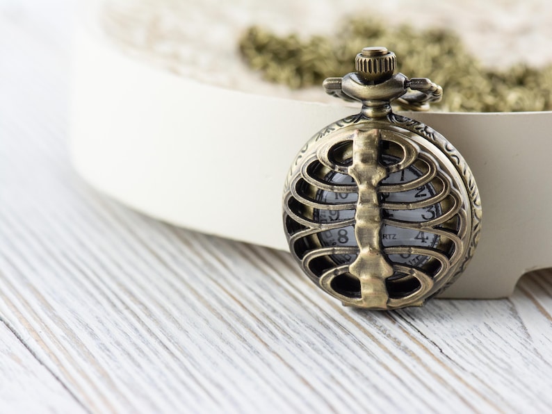 May include: A gold-toned pocket watch with a skeleton ribcage design. The watch face is visible through the ribs and has black numbers and hands.