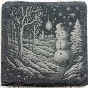 K&ouml;nnte beinhalten: Ein schwarzer Schieferuntersetzer mit einem wei&szlig;en, gravierten Design eines Schneemanns, der in einem schneebedeckten Wald mit einer Kirche im Hintergrund steht. Das Design enth&auml;lt auch Sterne und Schneeflocken.