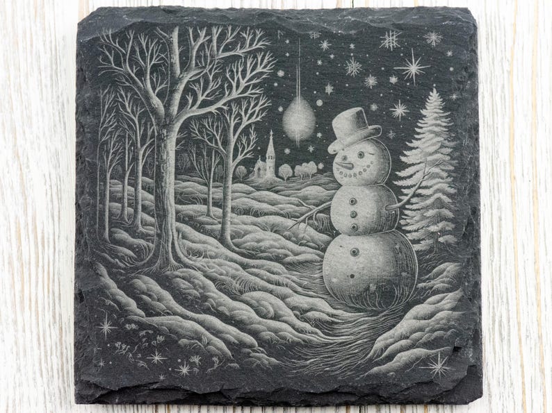 K&ouml;nnte beinhalten: Ein schwarzer Schieferuntersetzer mit einem wei&szlig;en, gravierten Design eines Schneemanns, der in einem schneebedeckten Wald mit einer Kirche im Hintergrund steht. Das Design enth&auml;lt auch Sterne und Schneeflocken.