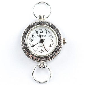May include: A silver Geneva quartz watch with a white face and black hands. The watch has rings at the top and bottom for attachment. The bezel is decorated with small, round, silver embellishments. The watch face displays the time with black numerals.