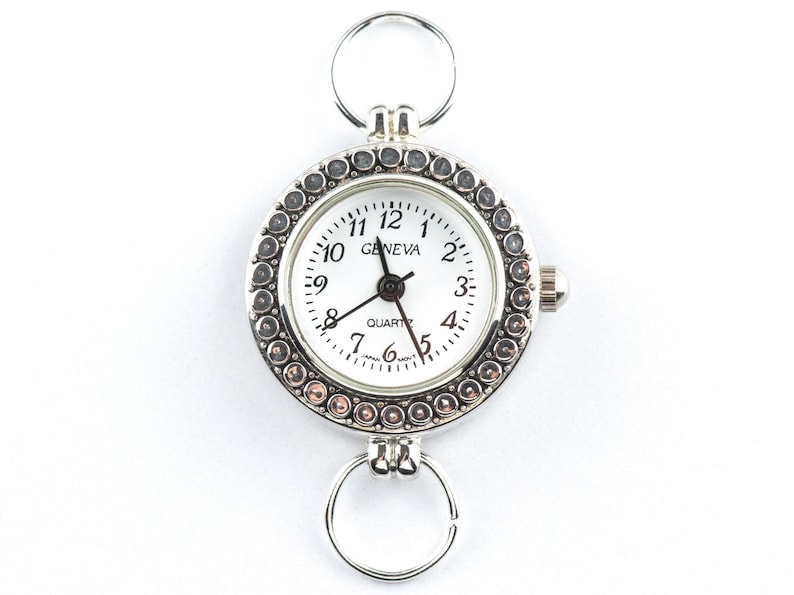 May include: A silver Geneva quartz watch with a white face and black hands. The watch has rings at the top and bottom for attachment. The bezel is decorated with small, round, silver embellishments. The watch face displays the time with black numerals.