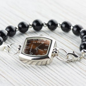 Puede incluir: Un reloj cuadrado plateado con una esfera marr&oacute;n y n&uacute;meros negros. El reloj est&aacute; unido a una pulsera de cuentas negras con cierres plateados.