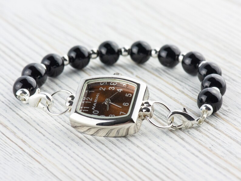 Puede incluir: Un reloj cuadrado plateado con una esfera marr&oacute;n y n&uacute;meros negros. El reloj est&aacute; unido a una pulsera de cuentas negras con cierres plateados.