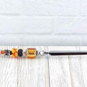 May include: A decorative letter opener with a silver blade and a beaded handle. The handle features orange, black, and yellow beads, along with a silver and amber-colored square bead. The opener is set against a white brick wall and a white wooden surface.