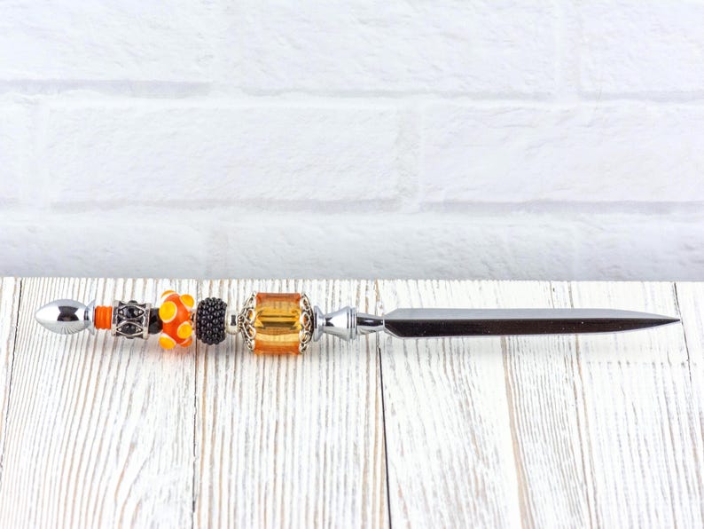 May include: A decorative letter opener with a silver blade and a beaded handle. The handle features orange, black, and yellow beads, along with a silver and amber-colored square bead. The opener is set against a white brick wall and a white wooden surface.
