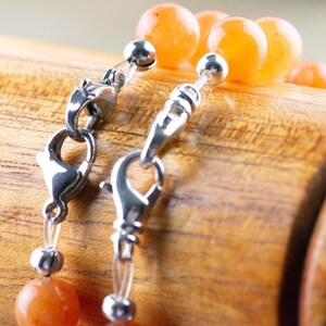 Op de afbeelding: Een close-up van een zilveren sluiting op een ketting met oranje carneol kralen. De ketting ligt op een houten oppervlak.