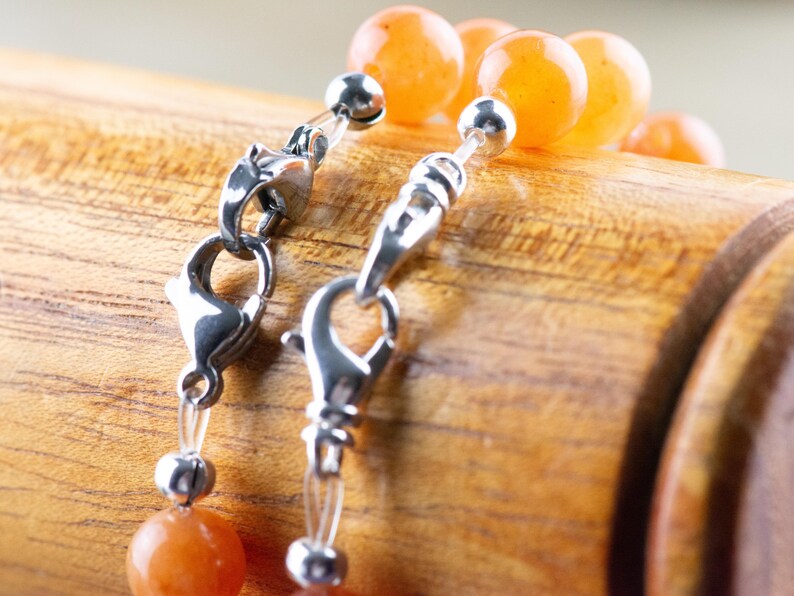 Op de afbeelding: Een close-up van een zilveren sluiting op een ketting met oranje carneol kralen. De ketting ligt op een houten oppervlak.