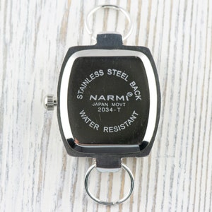 May include: A black and silver keychain watch with the text "STAINLESS STEEL BACK" and "WATER RESISTANT". The watch face is square with rounded edges and features the brand name "NARMI" and "JAPAN MOVT 2034-T".