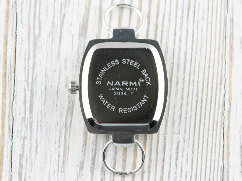 May include: A black and silver keychain watch with the text "STAINLESS STEEL BACK" and "WATER RESISTANT". The watch face is square with rounded edges and features the brand name "NARMI" and "JAPAN MOVT 2034-T".