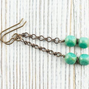 May include: A pair of handmade earrings featuring teal-colored, cube-shaped beads. The beads are connected by a chain to a bronze-colored ear hook. The earrings are displayed on a white wooden surface.
