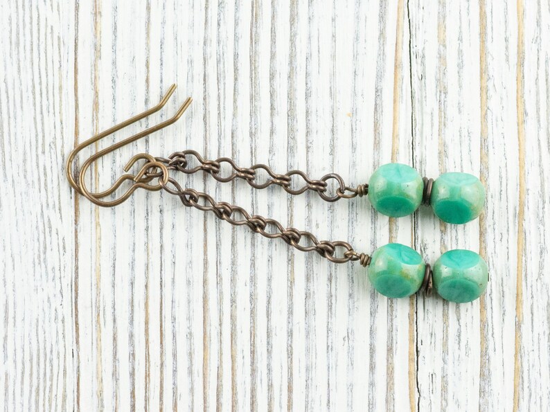 May include: A pair of handmade earrings featuring teal-colored, cube-shaped beads. The beads are connected by a chain to a bronze-colored ear hook. The earrings are displayed on a white wooden surface.