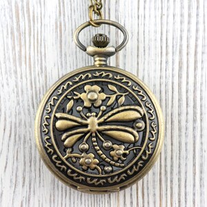 May include: Antique bronze pocket watch with a dragonfly design. The watch features a detailed dragonfly and floral pattern on the face, with a matching chain and clasp. The watch is round and has a decorative border.