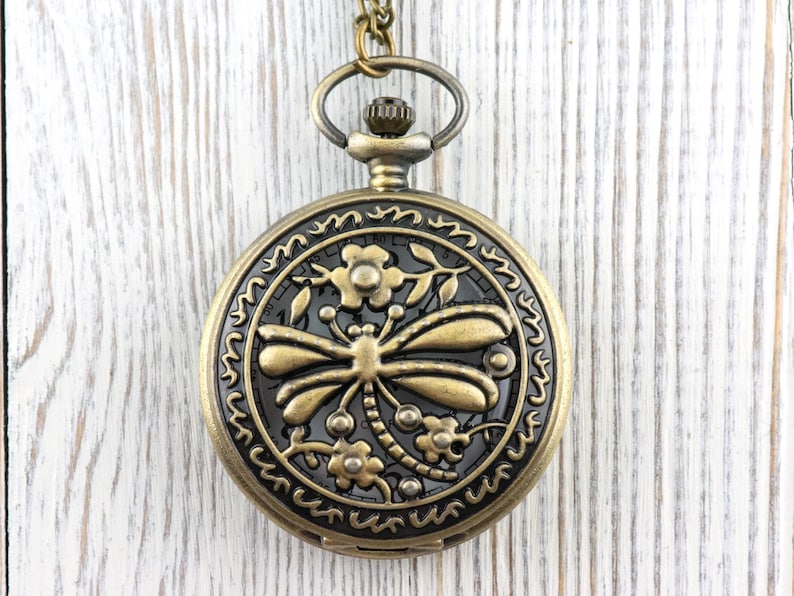 May include: Antique bronze pocket watch with a dragonfly design. The watch features a detailed dragonfly and floral pattern on the face, with a matching chain and clasp. The watch is round and has a decorative border.