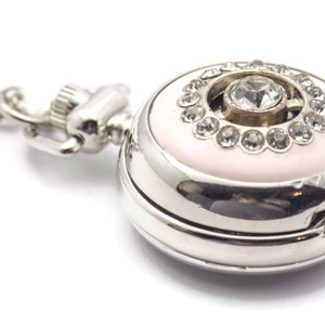 May include: A silver pocket watch with a pink face and a large rhinestone in the center surrounded by smaller rhinestones. The watch has a silver chain attached.