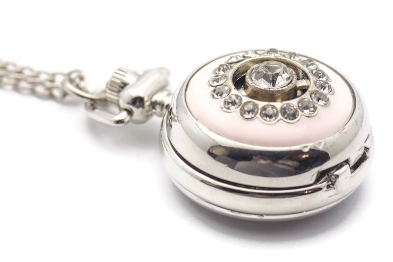 May include: A silver pocket watch with a pink face and a large rhinestone in the center surrounded by smaller rhinestones. The watch has a silver chain attached.