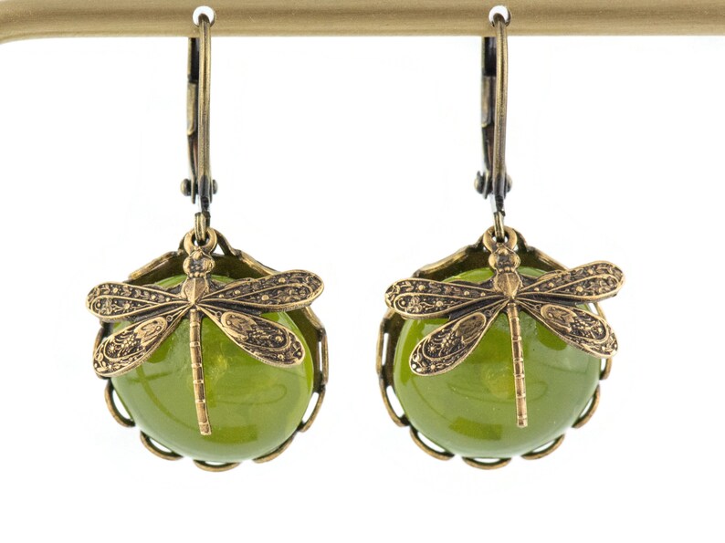 May include: A pair of green glass dangle earrings with a dragonfly charm. The earrings have a brass finish and a delicate design.