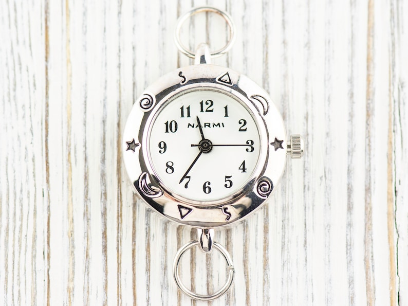 May include: A silver-toned round watch with a white face, black Arabic numerals, and the brand name NARMI printed in black. The bezel features engraved celestial symbols including stars, moons, and spirals. It includes two silver circular rings.