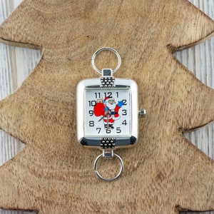Pu&ograve; includere: Un orologio da tasca quadrato argentato con un quadrante bianco e numeri neri. Il quadrante presenta un'immagine di cartone animato di Babbo Natale che tiene un sacco rosso. L'orologio ha una catena argentata attaccata alla parte superiore.