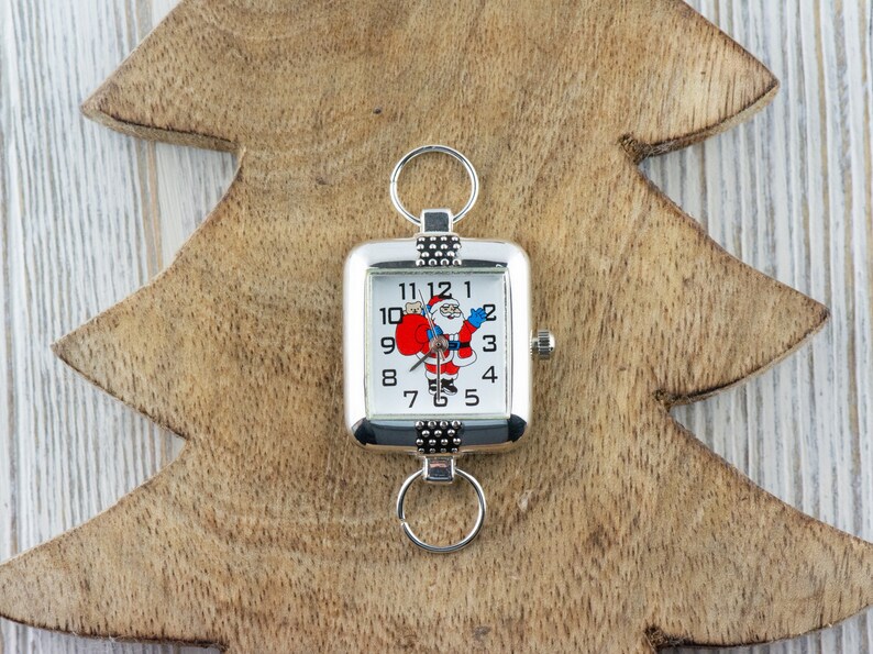 Pu&ograve; includere: Un orologio da tasca quadrato argentato con un quadrante bianco e numeri neri. Il quadrante presenta un'immagine di cartone animato di Babbo Natale che tiene un sacco rosso. L'orologio ha una catena argentata attaccata alla parte superiore.