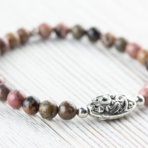 Puede incluir: Un brazalete de piedras preciosas rosa y marr&oacute;n con un dije de plata. El dije tiene un dise&ntilde;o floral.