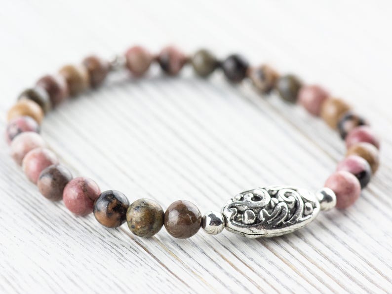 Puede incluir: Un brazalete de piedras preciosas rosa y marr&oacute;n con un dije de plata. El dije tiene un dise&ntilde;o floral.