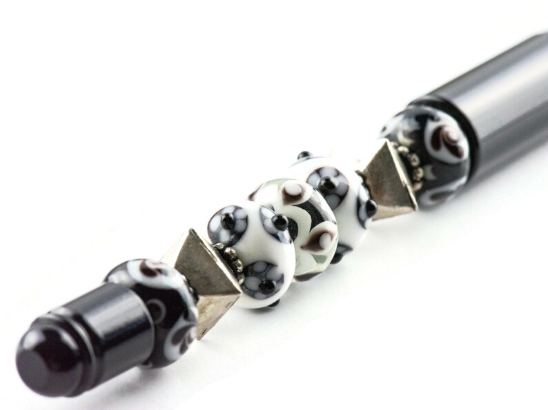 May include: A black pen with a silver and black patterned cap and a series of white and black glass beads with silver accents.