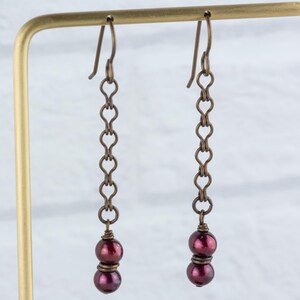 Puede incluir: Un par de pendientes colgantes de est&eacute;tica vintage. Cada pendiente presenta una cadena de peque&ntilde;os eslabones en forma de coraz&oacute;n, en tono bronce, que suspenden dos cuentas de color burdeos oscuro. Los pendientes se exhiben sobre un soporte dorado, frente a una pared de ladrillos blancos.