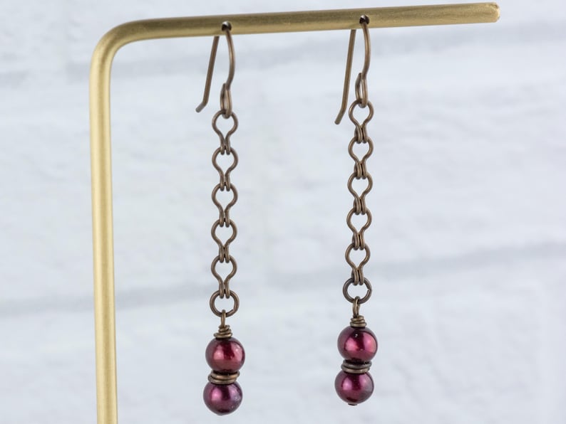 Puede incluir: Un par de pendientes colgantes de est&eacute;tica vintage. Cada pendiente presenta una cadena de peque&ntilde;os eslabones en forma de coraz&oacute;n, en tono bronce, que suspenden dos cuentas de color burdeos oscuro. Los pendientes se exhiben sobre un soporte dorado, frente a una pared de ladrillos blancos.