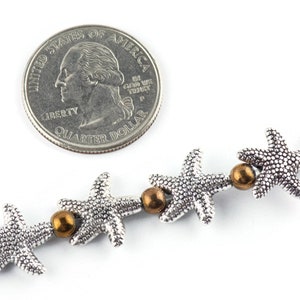 K&ouml;nnte beinhalten: Eine silberne Kette mit silbernen Seestern-Anh&auml;ngern und bronzenen Perlen. Die Kette wird neben einem US-amerikanischen Viertel-Dollar zur Gr&ouml;&szlig;enreferenz gezeigt.