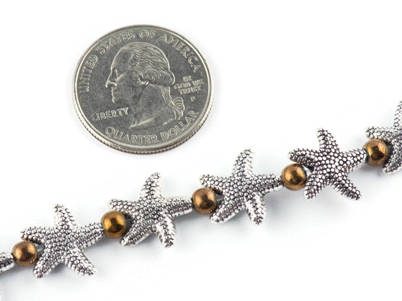 K&ouml;nnte beinhalten: Eine silberne Kette mit silbernen Seestern-Anh&auml;ngern und bronzenen Perlen. Die Kette wird neben einem US-amerikanischen Viertel-Dollar zur Gr&ouml;&szlig;enreferenz gezeigt.