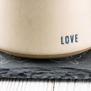 Puede incluir: Una taza de cer&aacute;mica blanca con la palabra "LOVE" en letras azules en el lateral. La taza est&aacute; sobre un posavasos de pizarra negra con un dise&ntilde;o decorativo.