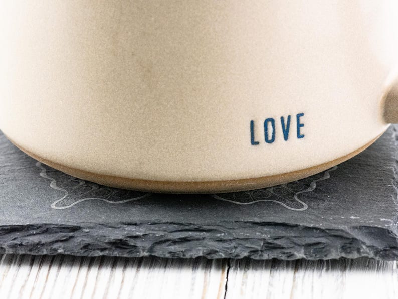 Puede incluir: Una taza de cer&aacute;mica blanca con la palabra "LOVE" en letras azules en el lateral. La taza est&aacute; sobre un posavasos de pizarra negra con un dise&ntilde;o decorativo.