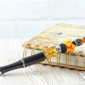 May include: A decorative pen with a black barrel and silver accents. The pen features a series of colorful beads, including orange, yellow, and black, and is topped with a black finial. The pen rests on a spiral-bound notebook.