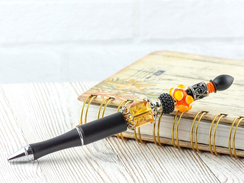 May include: A decorative pen with a black barrel and silver accents. The pen features a series of colorful beads, including orange, yellow, and black, and is topped with a black finial. The pen rests on a spiral-bound notebook.