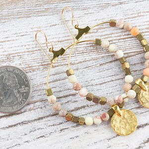 May include: Gold hoop earrings with a hammered gold disc charm and a mix of beige, white, and peach colored beads.
