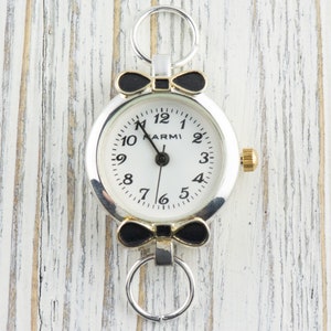 May include: A silver and gold watch with a white face and black bow accents. The watch has black numbers and a black hand. The watch is on a white wooden background.