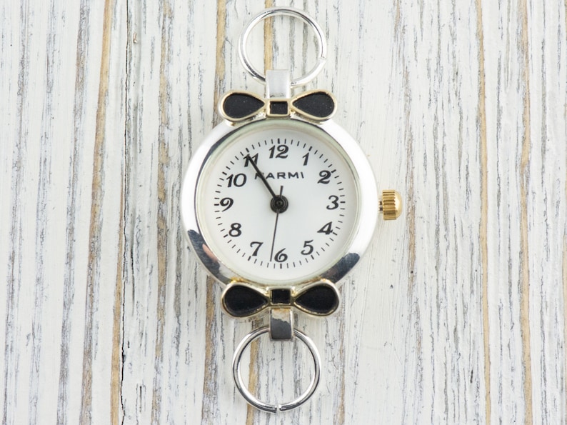 May include: A silver and gold watch with a white face and black bow accents. The watch has black numbers and a black hand. The watch is on a white wooden background.