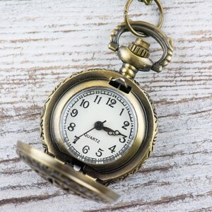 May include: A gold-toned pocket watch with an open face and a white dial. The watch has black hands and black Roman numeral hour markers. The watch face also has the word "QUARTZ" printed on it.
