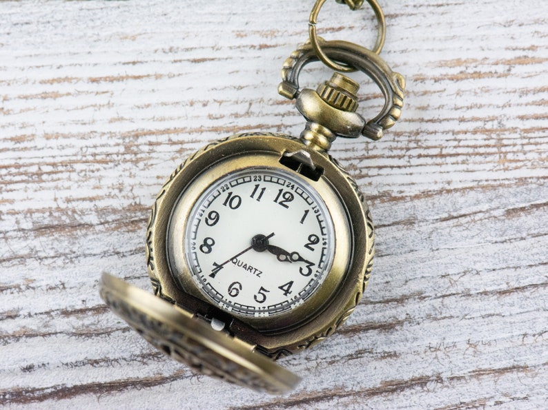 May include: A gold-toned pocket watch with an open face and a white dial. The watch has black hands and black Roman numeral hour markers. The watch face also has the word "QUARTZ" printed on it.