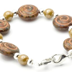 May include: A bracelet with brown, oval beads with an orange and brown spiral pattern. The bracelet also has olive-green beads, silver-coloured spacers and a clasp. The jewellery is set against a white background.