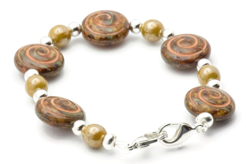 May include: A bracelet with brown, oval beads with an orange and brown spiral pattern. The bracelet also has olive-green beads, silver-coloured spacers and a clasp. The jewellery is set against a white background.