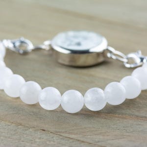 May include: A white beaded bracelet with a silver watch attached. The bracelet is made of round, white beads and the watch has a silver face and hands.