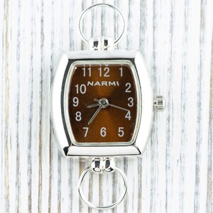 Puede incluir: Un reloj cuadrado plateado con una esfera marr&oacute;n y agujas negras. El reloj tiene una cadena plateada unida a la parte superior y una perilla plateada en el lateral. La esfera del reloj tiene la palabra "NARMI" impresa en ella.