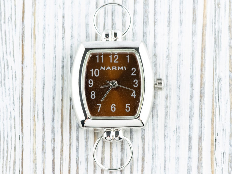 Puede incluir: Un reloj cuadrado plateado con una esfera marr&oacute;n y agujas negras. El reloj tiene una cadena plateada unida a la parte superior y una perilla plateada en el lateral. La esfera del reloj tiene la palabra "NARMI" impresa en ella.